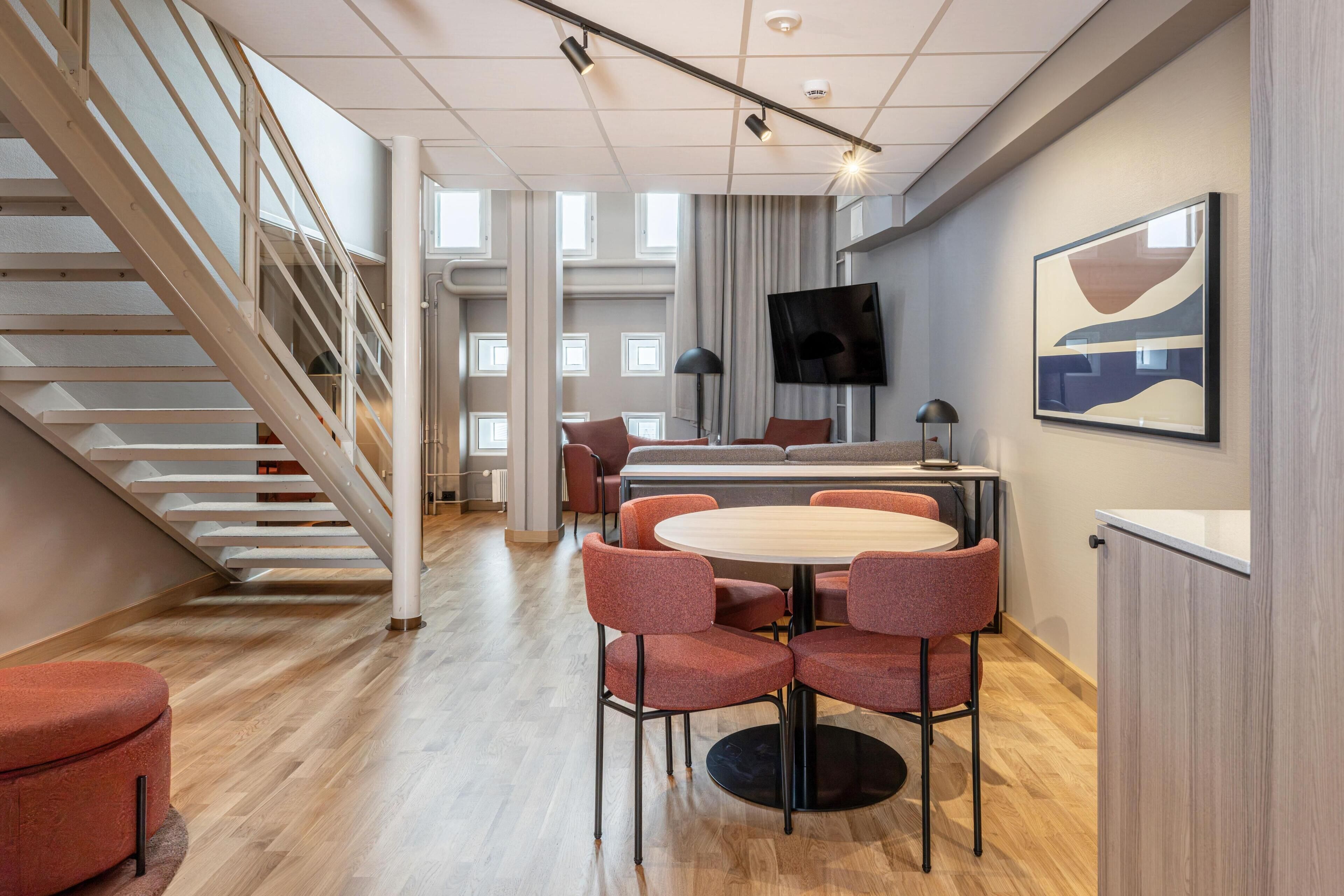 A modern hotel room with a staircase, a dining area with round table and chairs, a flat-screen TV mounted on the wall, and a large abstract painting.
