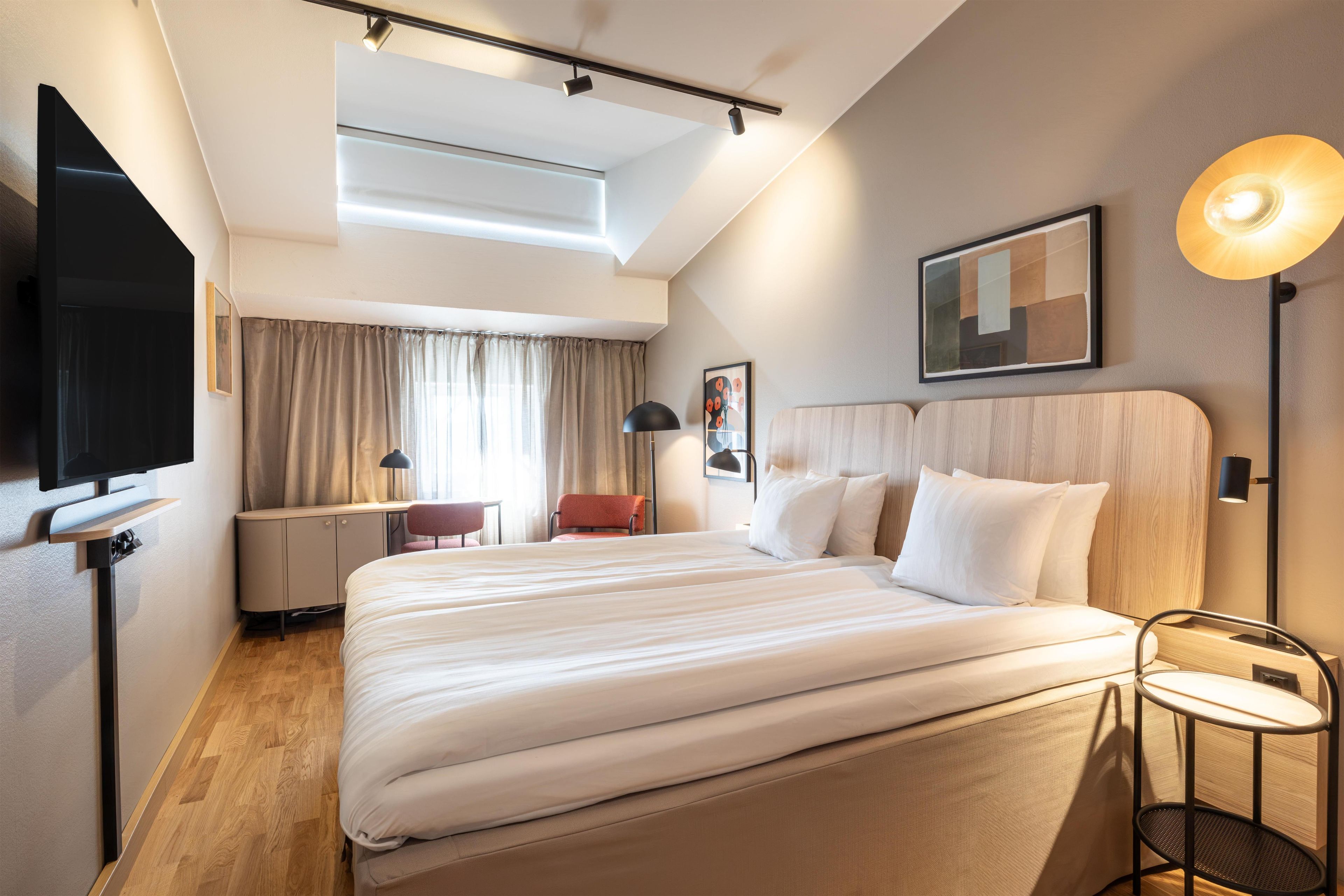 A hotel room with a bed, a desk, a chair, a TV, and a lamp.