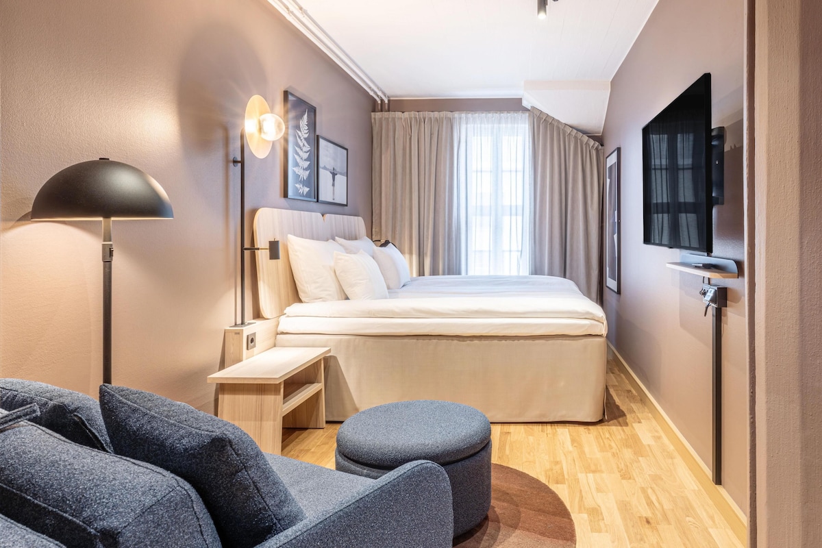A modern hotel room with a bed, a desk, a chair, a TV, and a window with curtains.