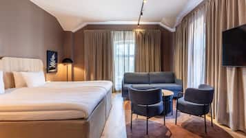 Habitación de hotel con cama, sofá, dos sillones, una mesita, televisor de pantalla plana y ventana con cortinas.
