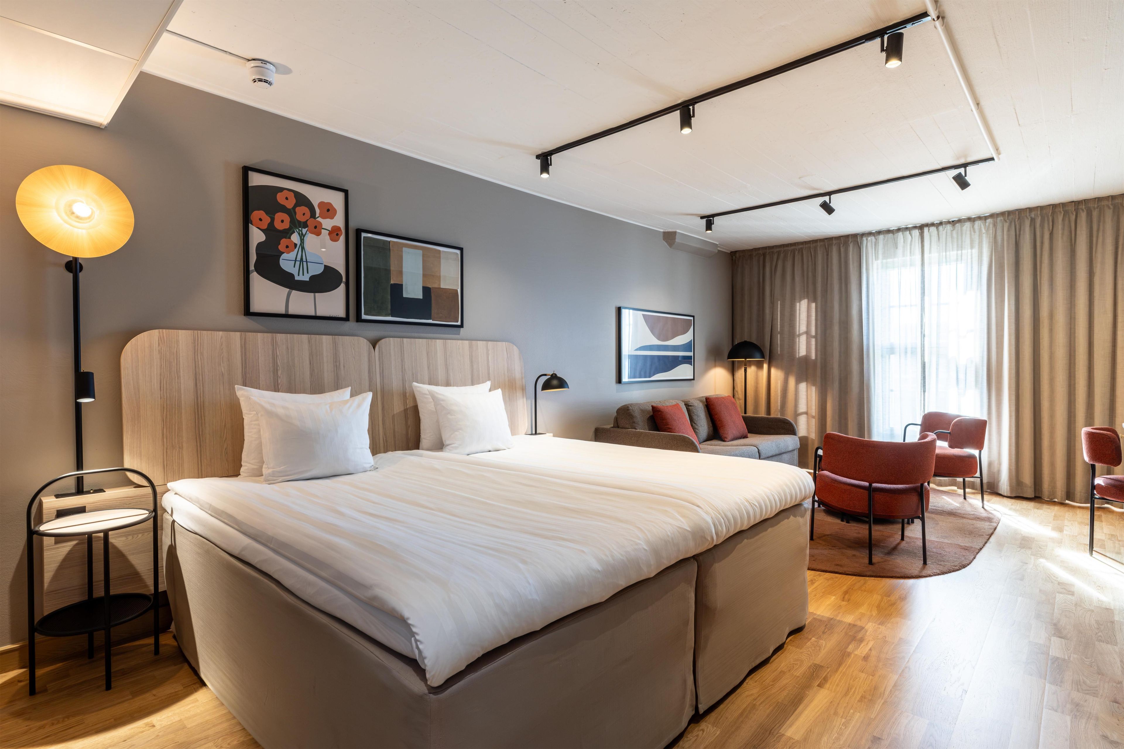 A modern hotel room with a large bed, a desk, a chair, and a sofa. There are two paintings on the wall and a ceiling light.