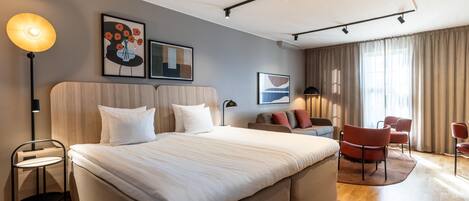 A modern hotel room with a large bed, a desk, a chair, and a sofa. There are two paintings on the wall and a ceiling light.