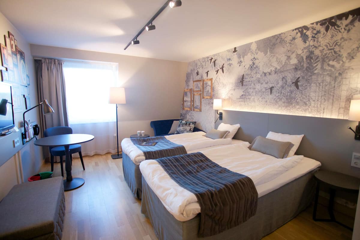 A hotel room with two beds, a desk, and a chair. There are wall-mounted lamps and a large window with curtains.