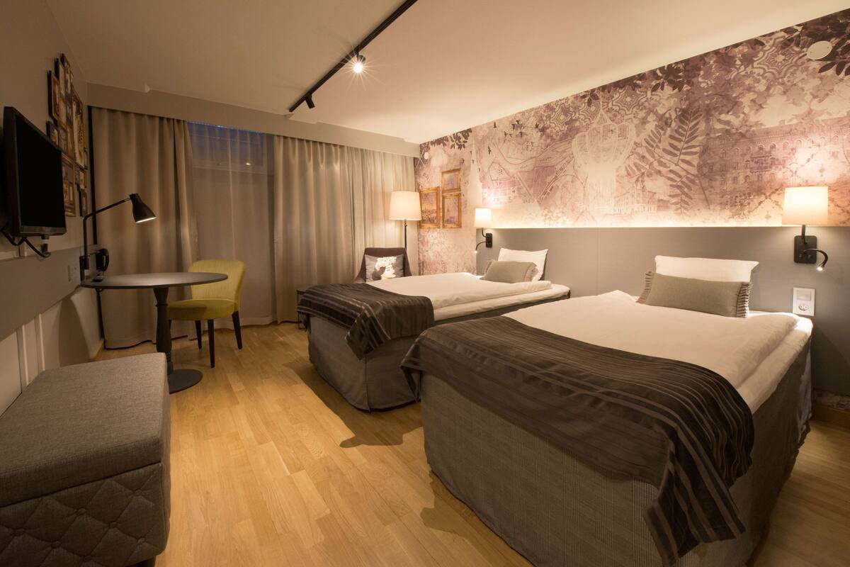 A hotel room with a bed, two bedside tables with lamps, a small round table with a chair, a sofa, and a large wall tapestry.