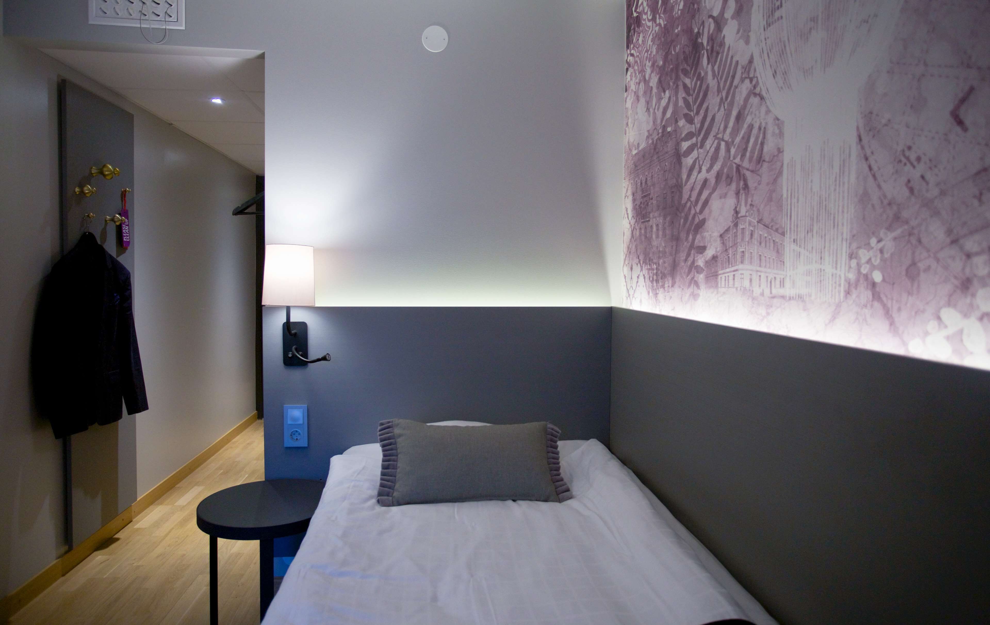 Een hotelkamer met een bed, een klein tafeltje, een wandlamp en een kapstok met een jas.