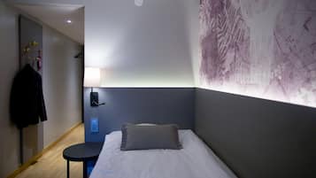 Une chambre d’hôtel avec un lit, une petite table, une lampe fixée au mur et un porte-manteau sur lequel est accrochée une veste.