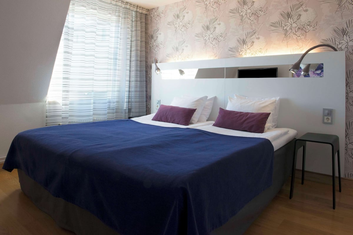A modern bedroom with a large bed, a nightstand, a wall-mounted lamp, and a window with curtains.