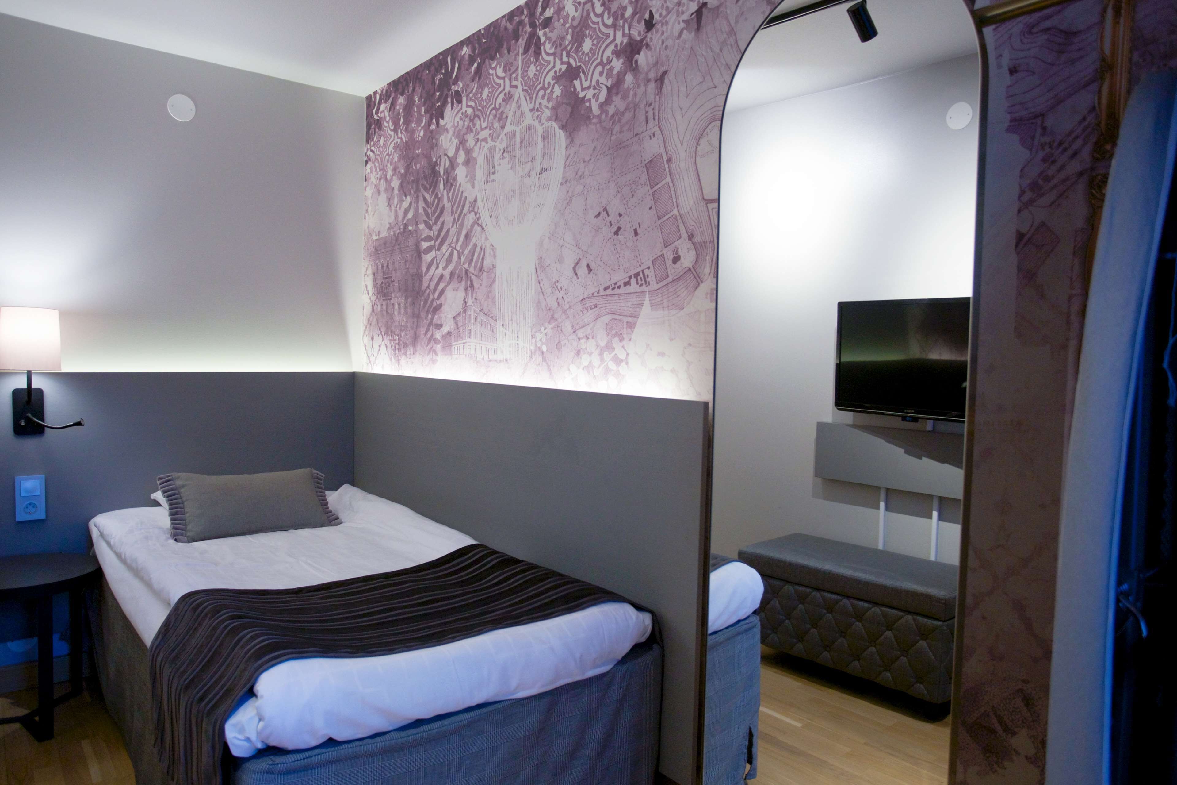 A modern hotel room with a bed, a desk, a chair, a TV, and a large mirror.