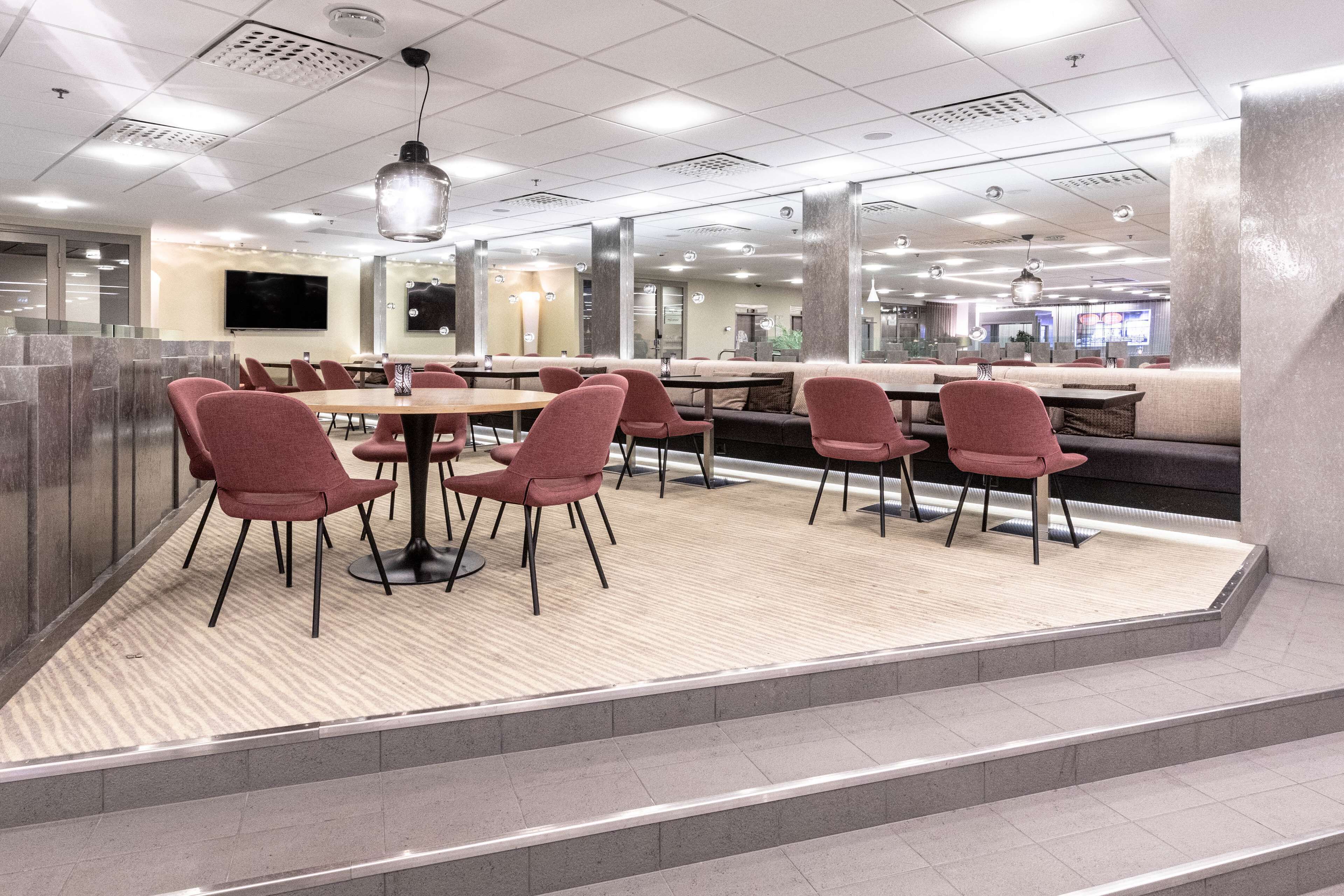 Een moderne eetruimte met hoge tafels en rode stoelen, een groot scherm en een zithoek met een bank en extra stoelen.
