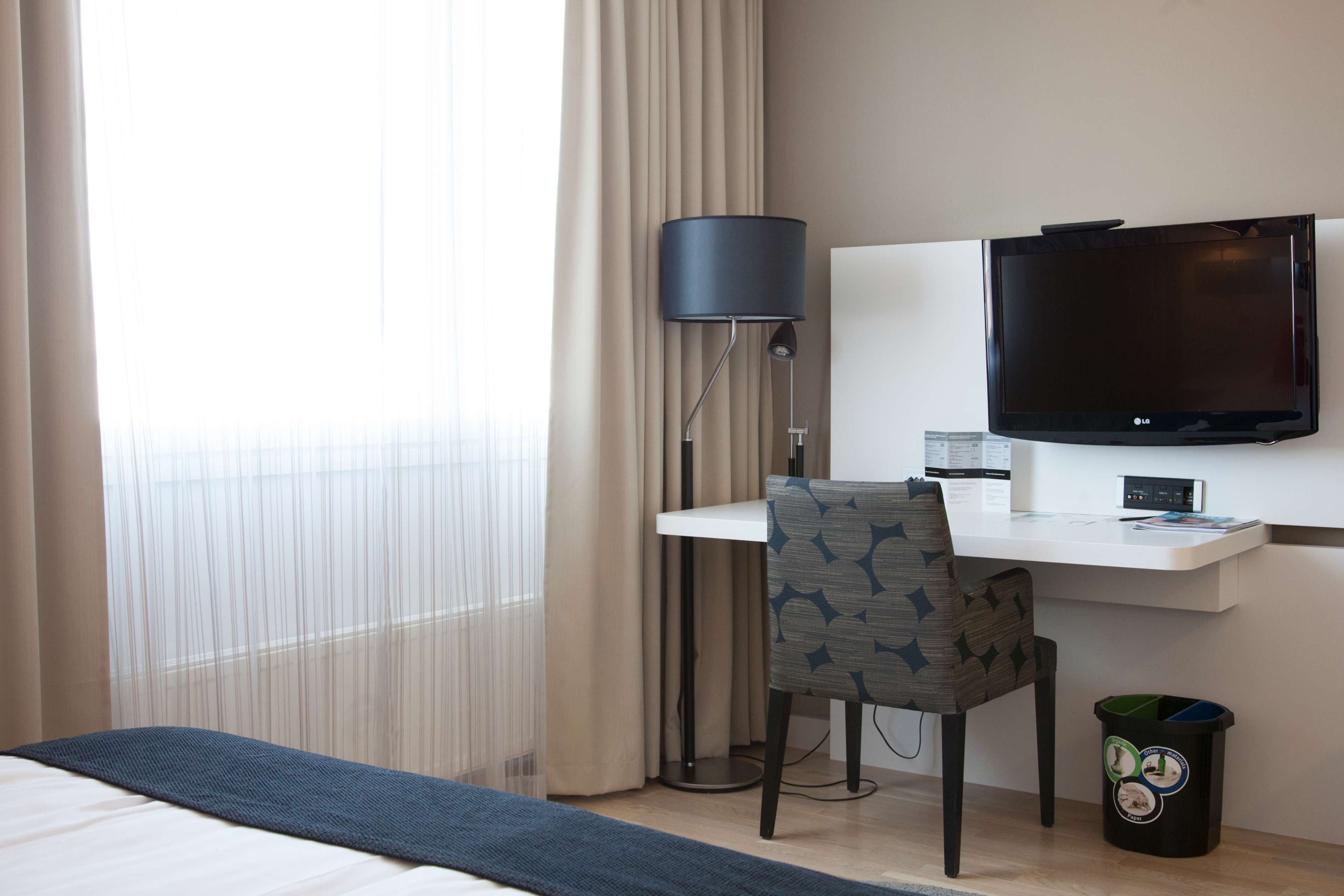 A hotel room with a bed, a desk with a chair, a television, a lamp, and a trash bin.