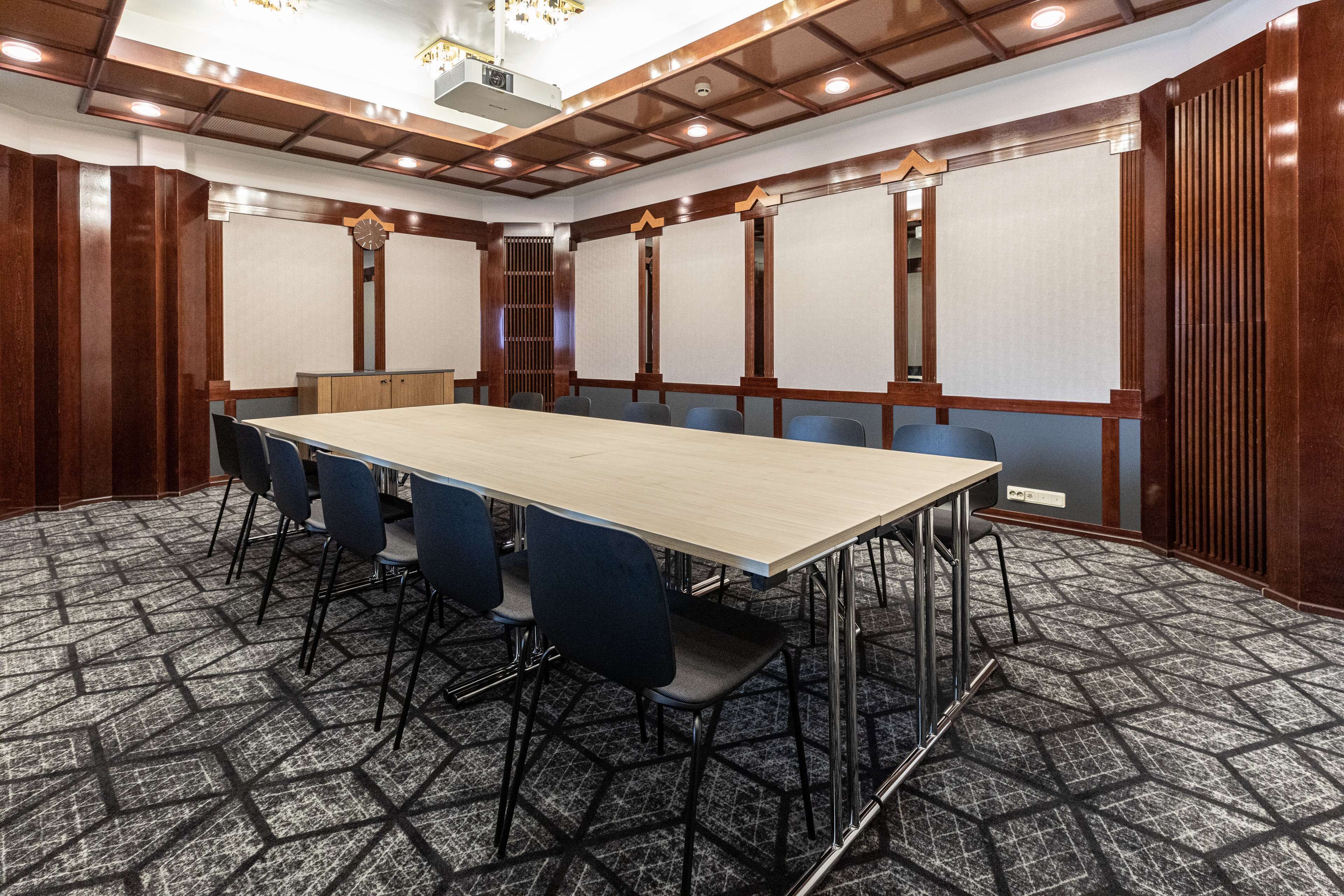 A conference room with a long table, blue chairs, a clock, and a patterned carpet.