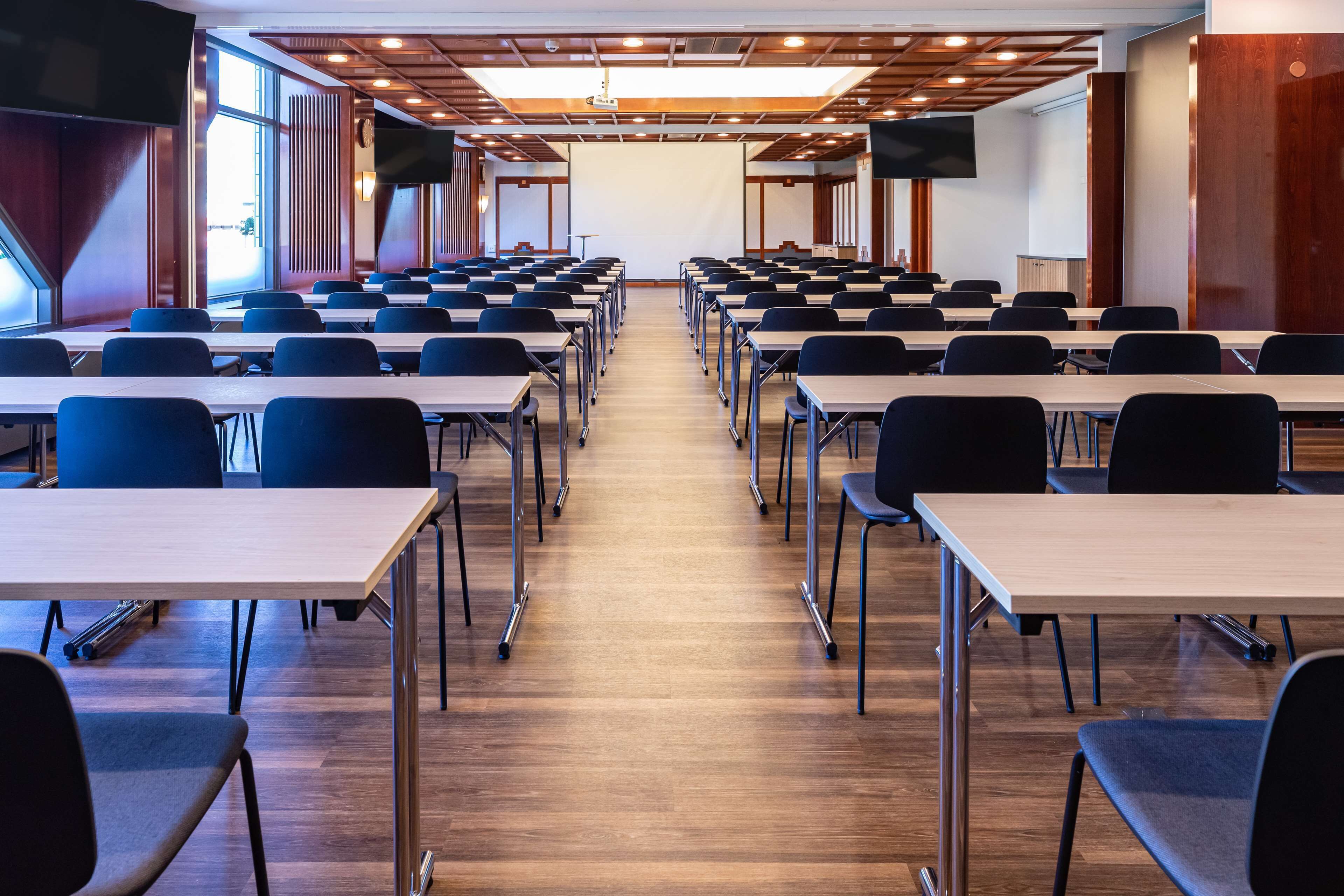 Une salle de conférence avec des rangées de tables et de chaises, un écran de projection et une grande fenêtre.