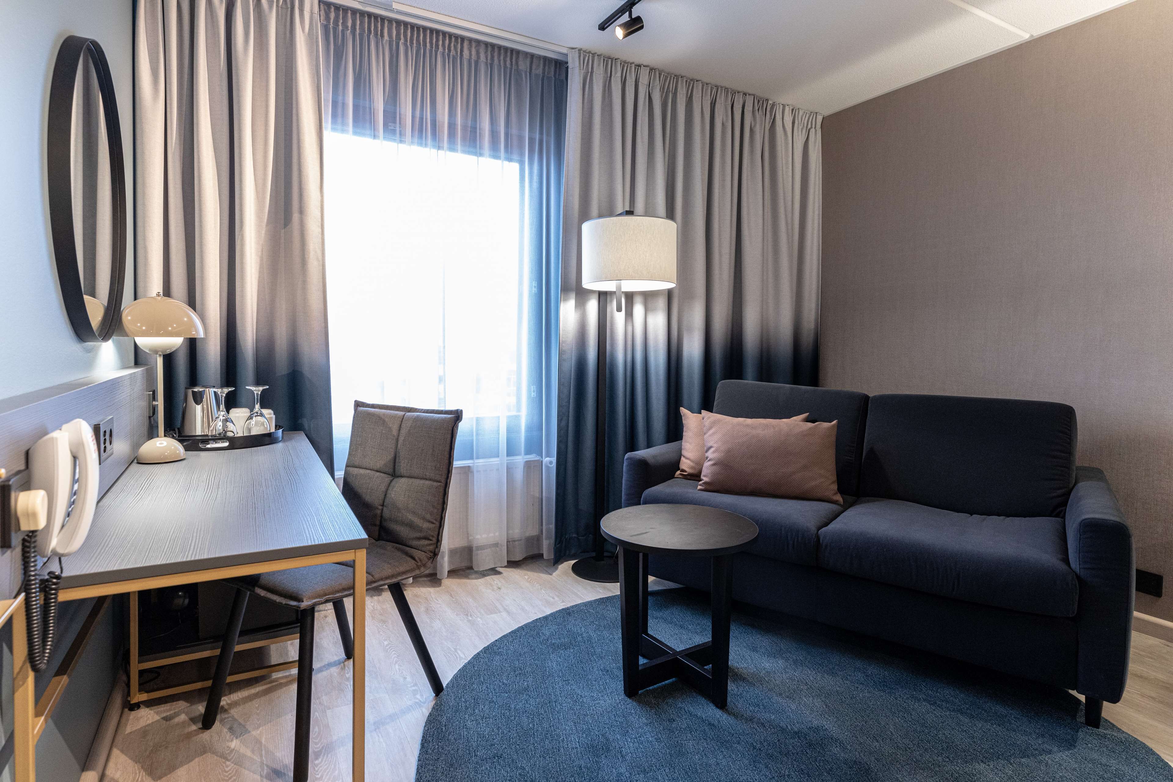 A modern hotel room with a dark sofa, a round coffee table, a desk with a telephone, and a large window with curtains.