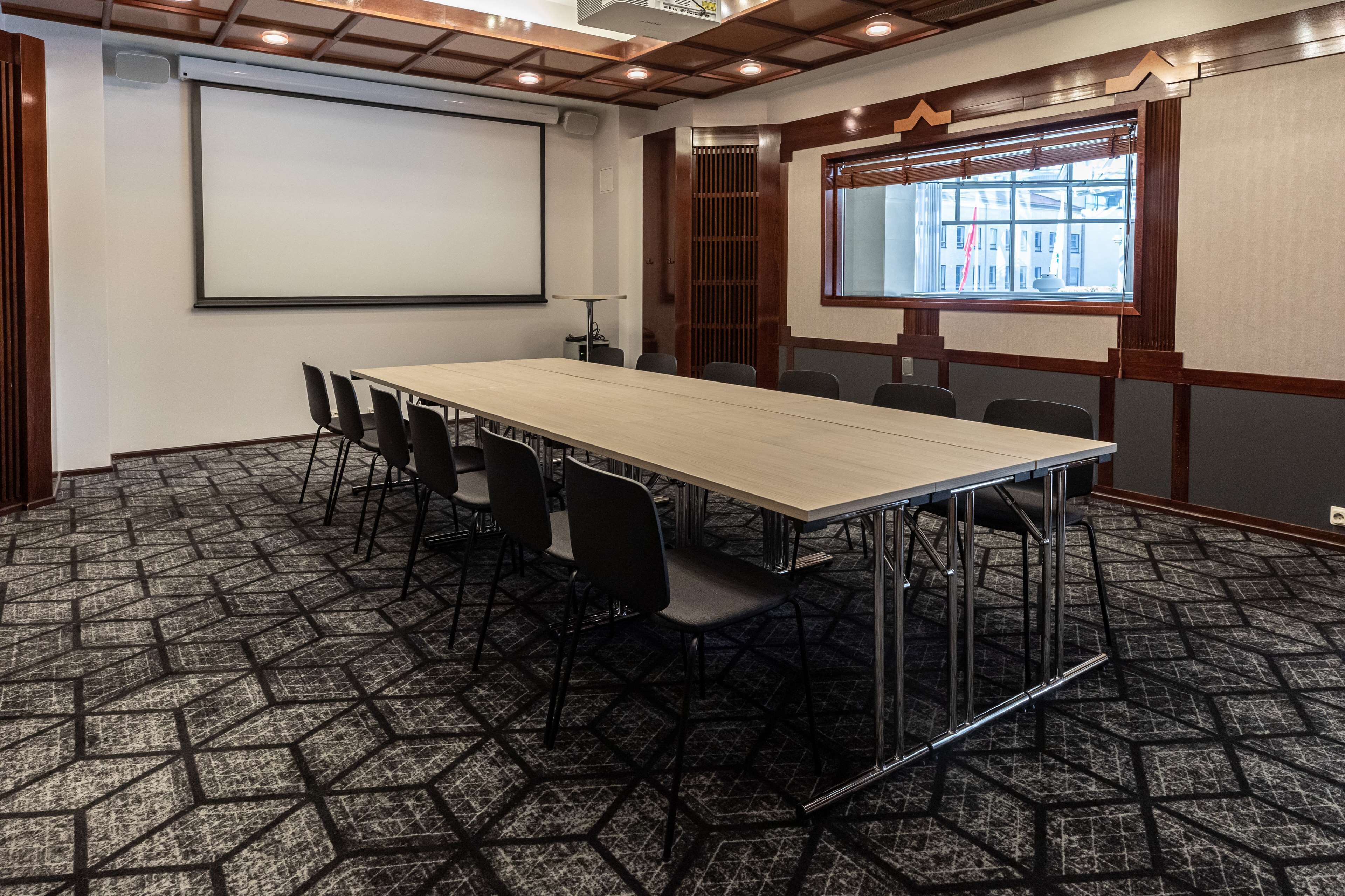 A conference room with a long table, chairs, a projector screen, and a window with a view of buildings.