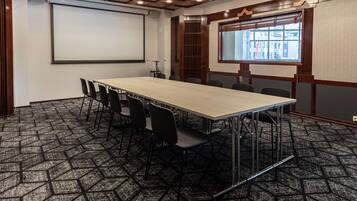 A conference room with a long table, chairs, a projector screen, and a window with a view of buildings.