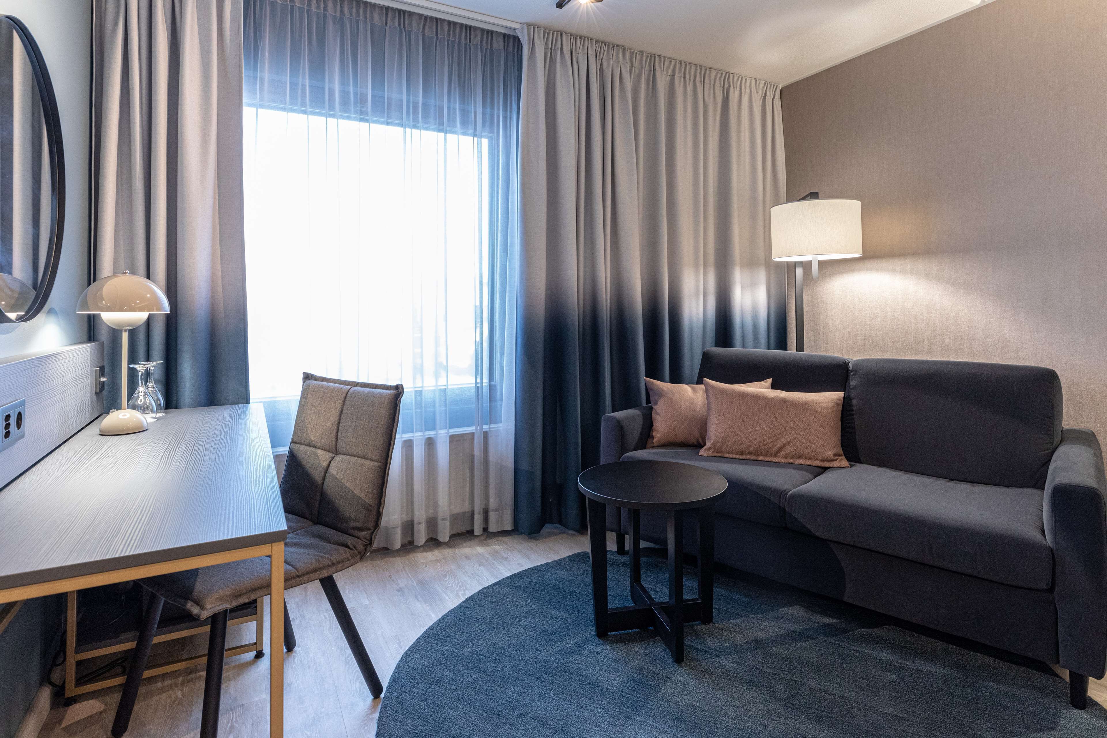 A modern hotel room with a dark grey sofa, a round coffee table, a dining table with chairs, and a large window with curtains.