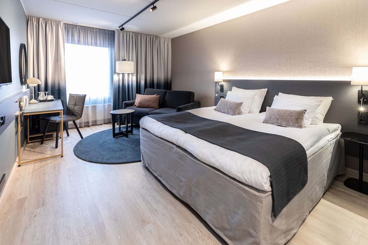 A modern hotel room with a large bed, a desk, a chair, and a small table.