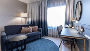 Une chambre d’hôtel moderne avec un canapé gris, une petite table ronde, une table à manger avec des chaises, une lampe, un miroir et une fenêtre avec des rideaux.