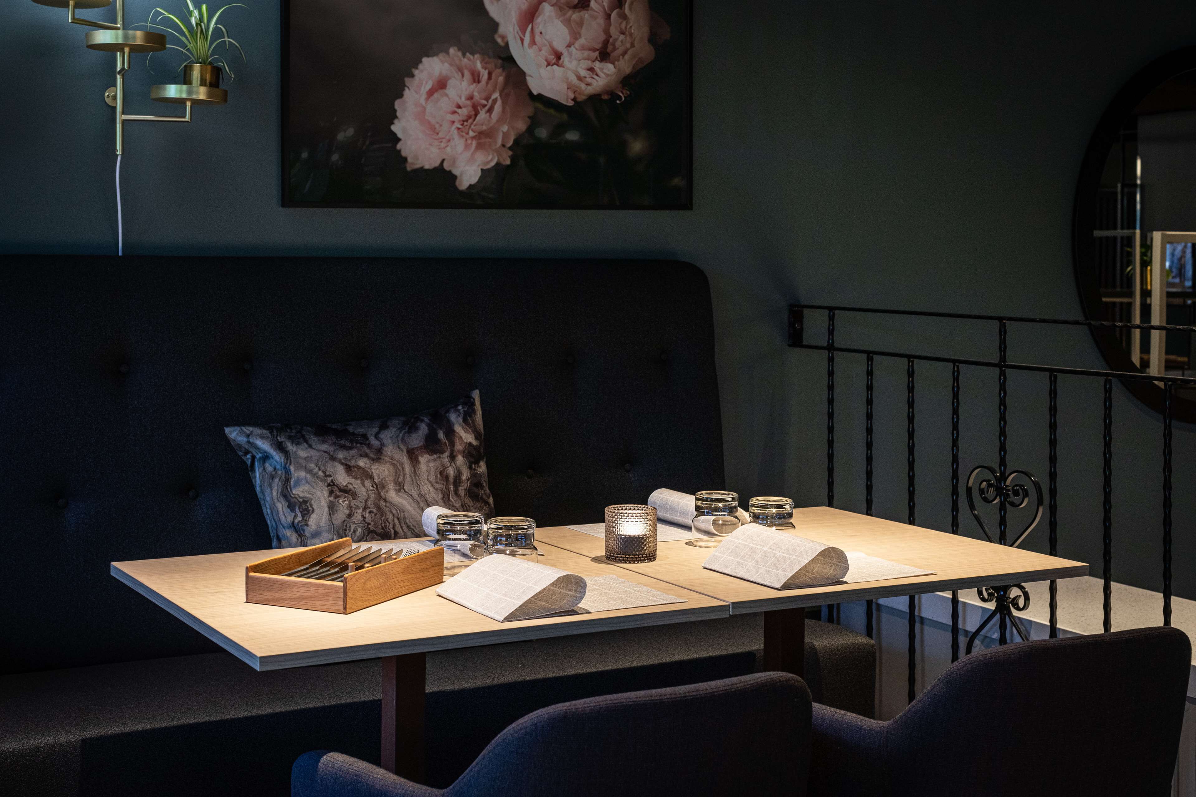 Un comedor con una mesa dispuesta para dos personas, que incluye una caja de cubiertos, dos velas y menús. Un sofá cómodo y una obra de arte floral enmarcada se encuentran al fondo.