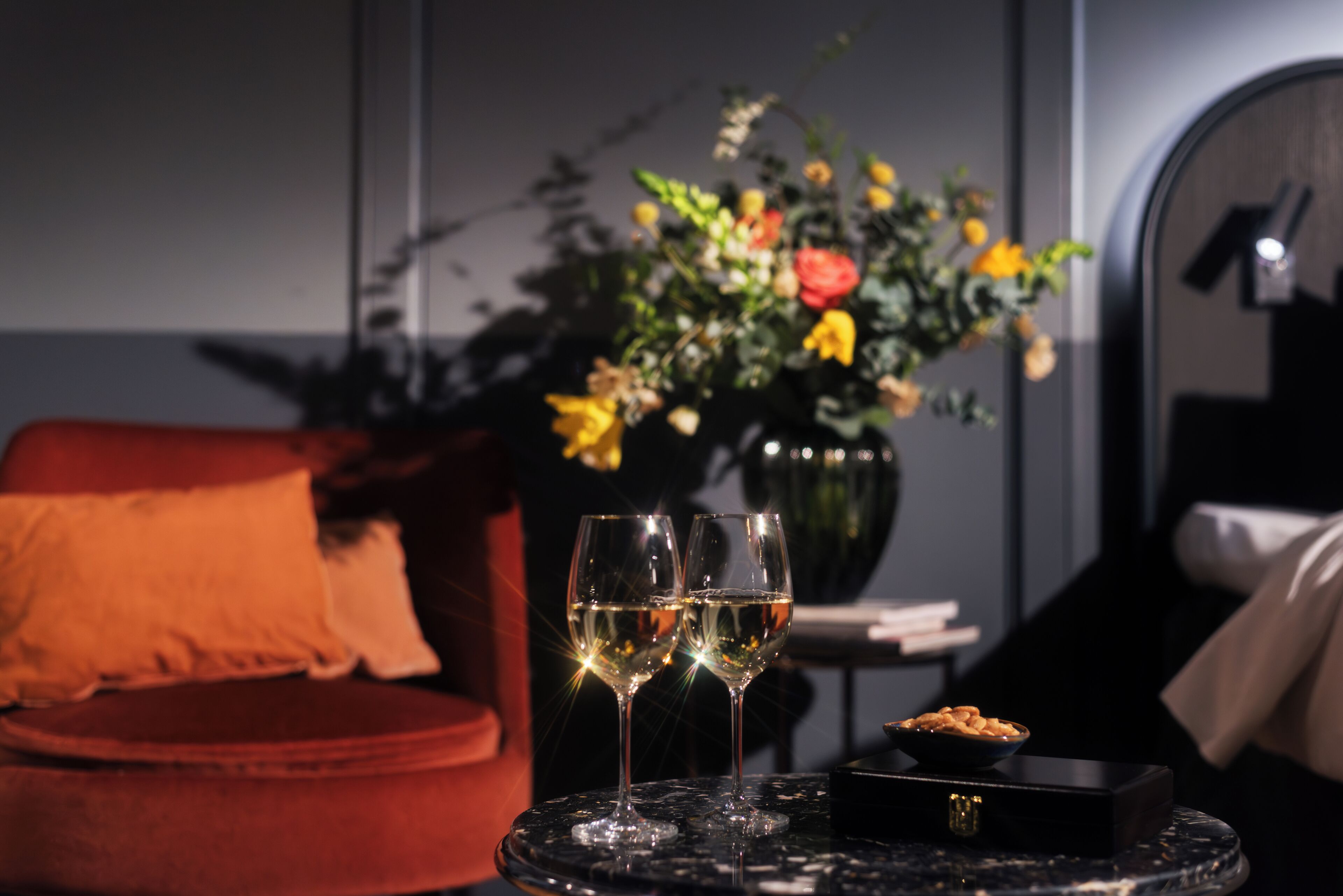 Una habitación con un sofá rojo, una mesita con dos copas de vino blanco, un jarrón con flores y una caja negra.