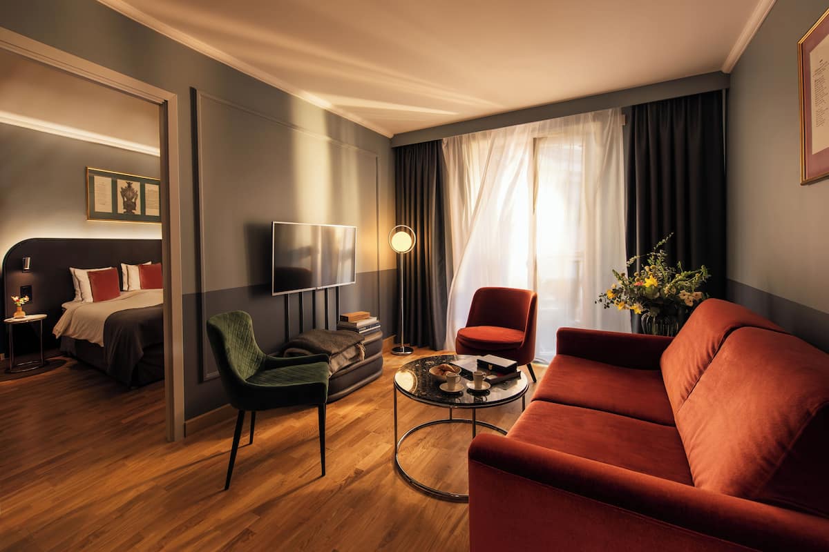 A modern hotel room with a wooden floor, a red sofa, a green armchair, a small round table, and a bed with a dark headboard.