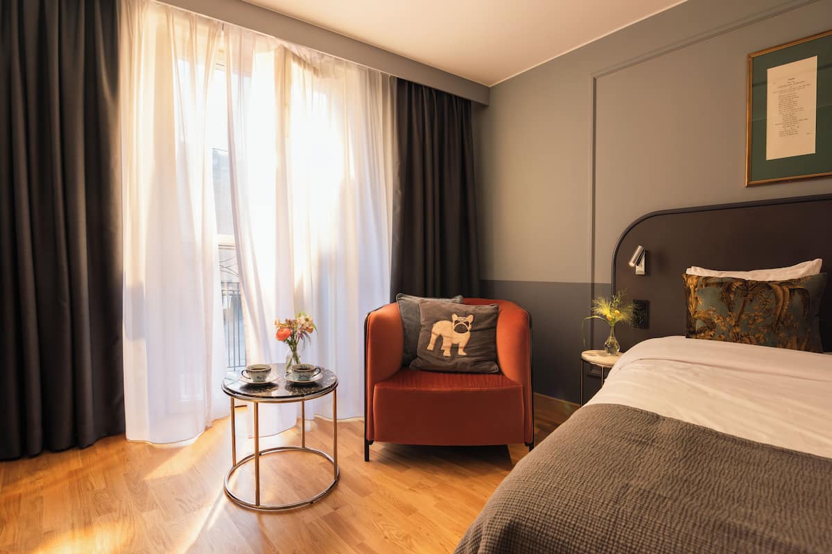 A hotel room with a bed, a chair with a cushion, a small table with a vase, and a window with curtains.