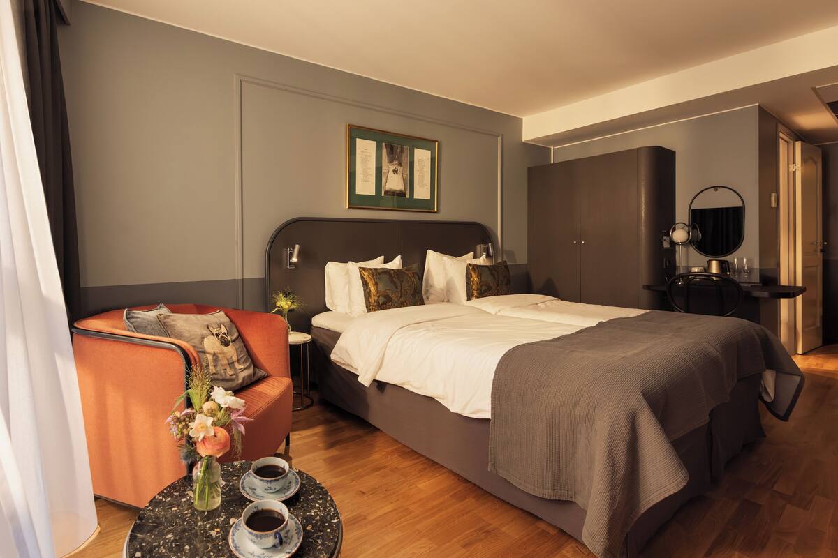 A modern hotel room with a large bed, a seating area with a table, and a wardrobe.