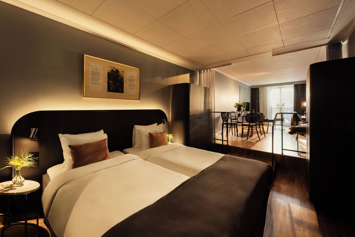 A modern hotel room with a large bed, a small side table with a lamp, a framed picture on the wall, and a view of the dining area.