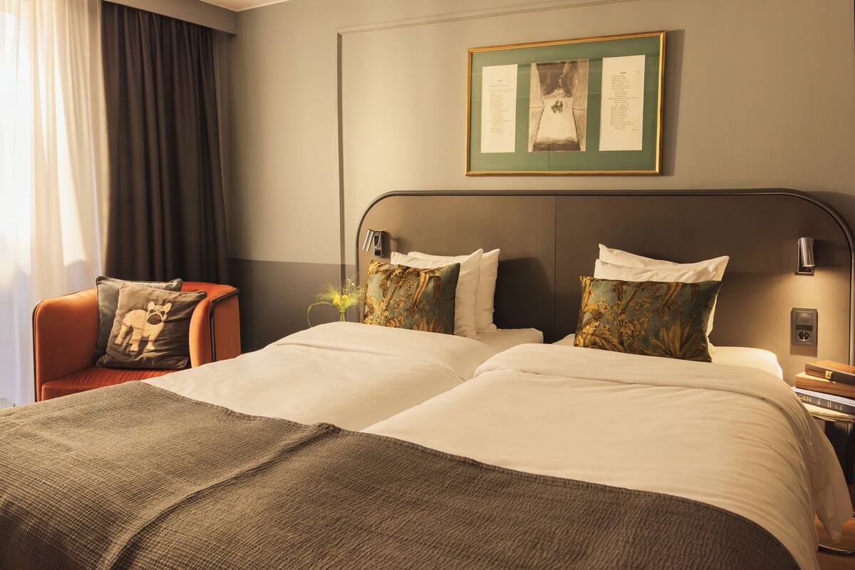 A hotel room with a large bed, two pillows, a nightstand, a lamp, a framed picture on the wall, and a chair with a cushion.