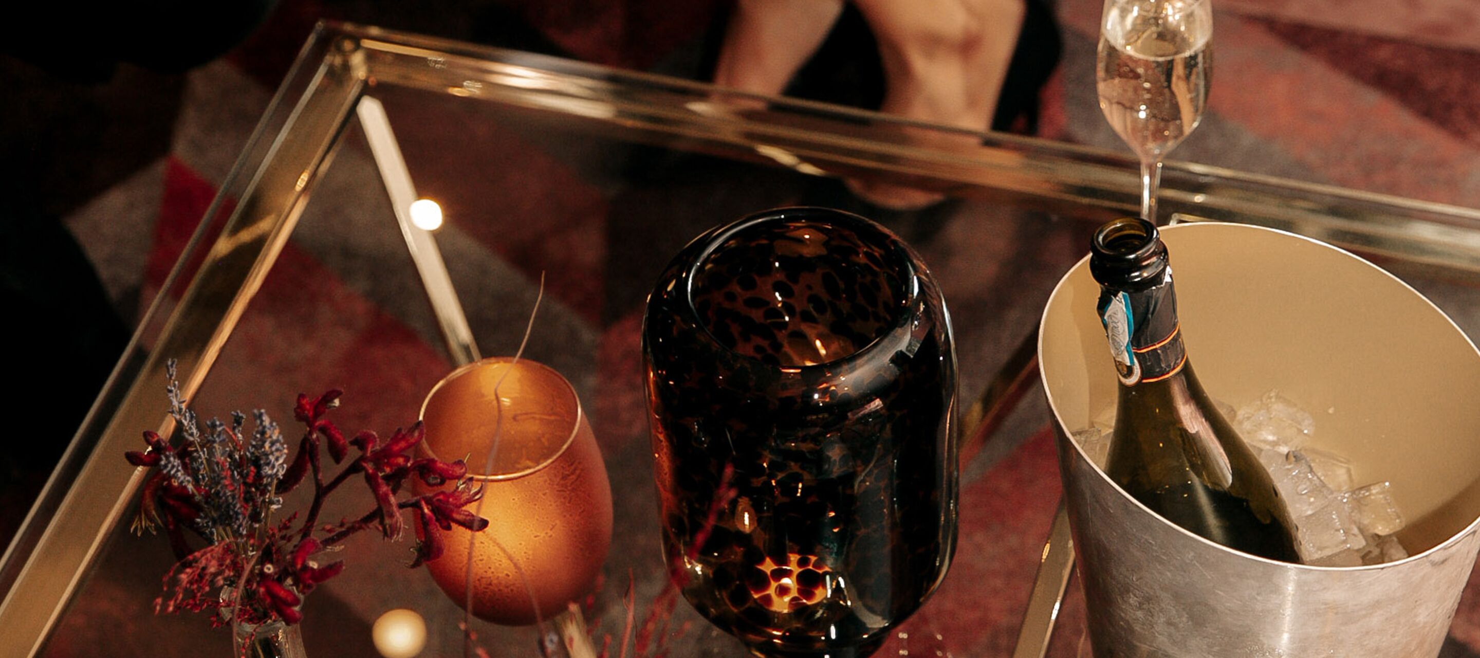 A glass table with a bottle of champagne, a glass of red wine, and a vase with dried flowers.