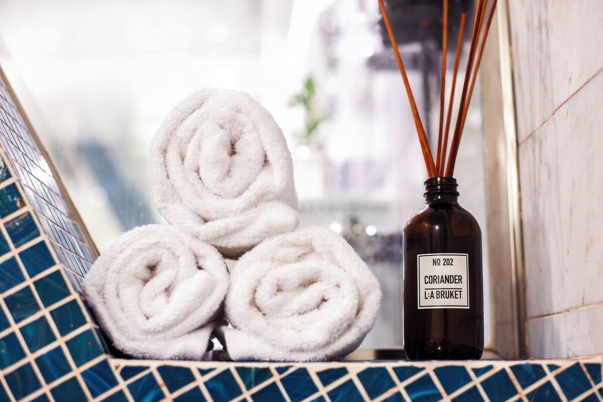 A bottle of Coriander L'A Bruket with reed diffusers, placed on a tiled surface next to rolled towels.
