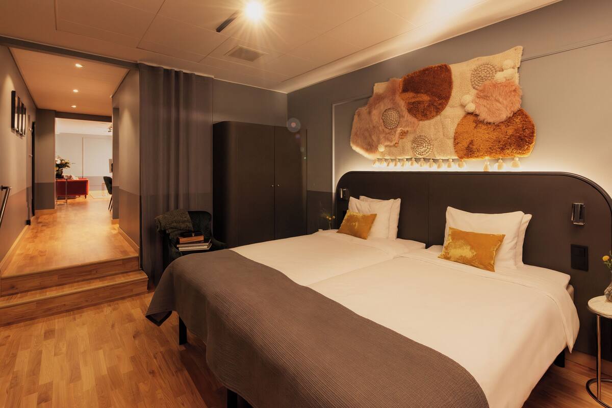 A modern hotel room with a large bed, a dark headboard, and a decorative wall hanging.
