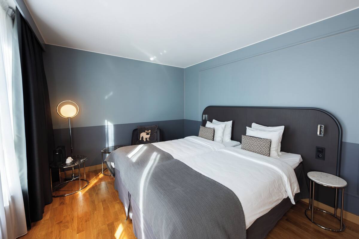 A modern hotel room with a large bed, a bedside table, a lamp, and a small stool.