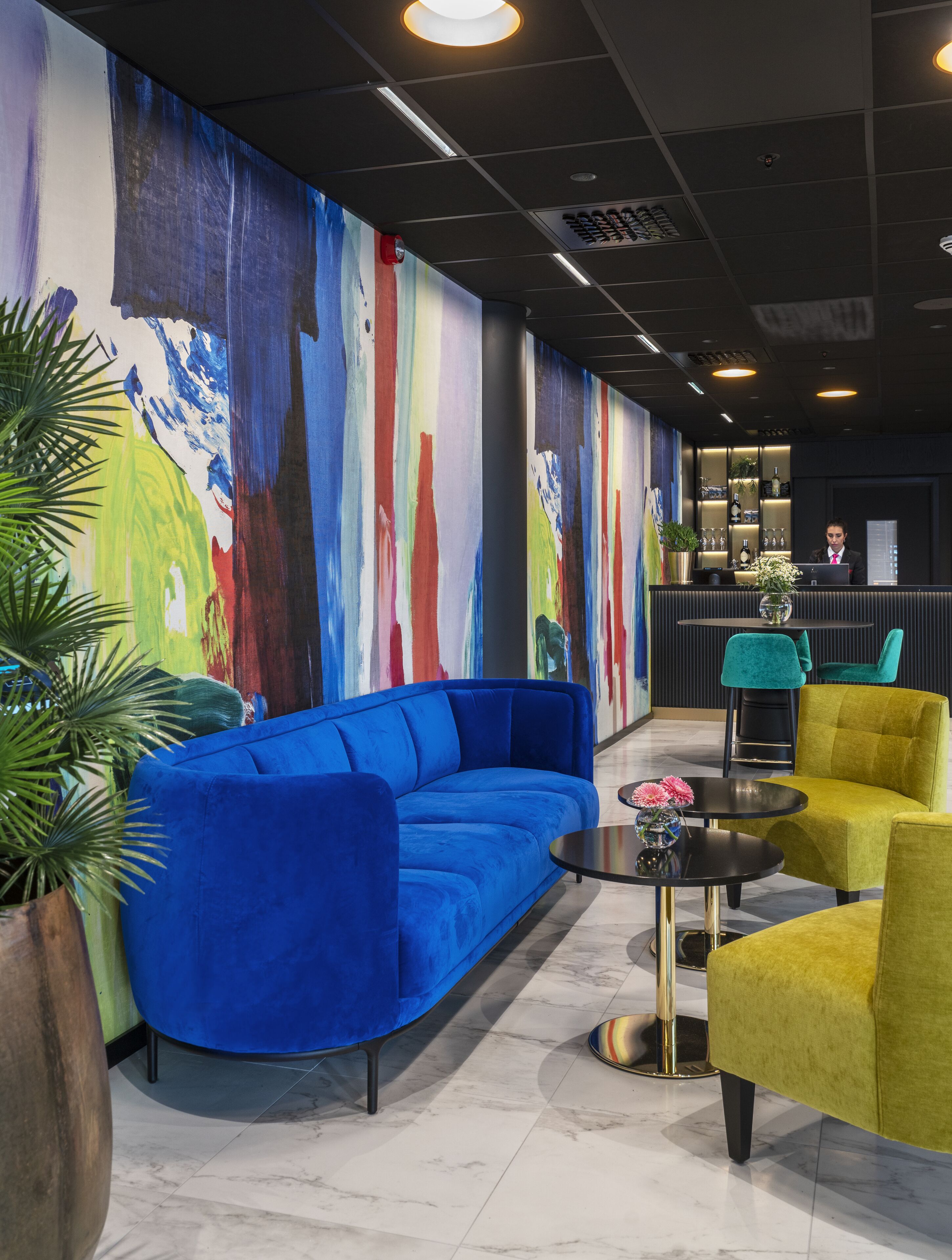 A modern lobby with a blue sofa, yellow armchairs, a round table with flowers, and a colorful abstract mural on the wall.