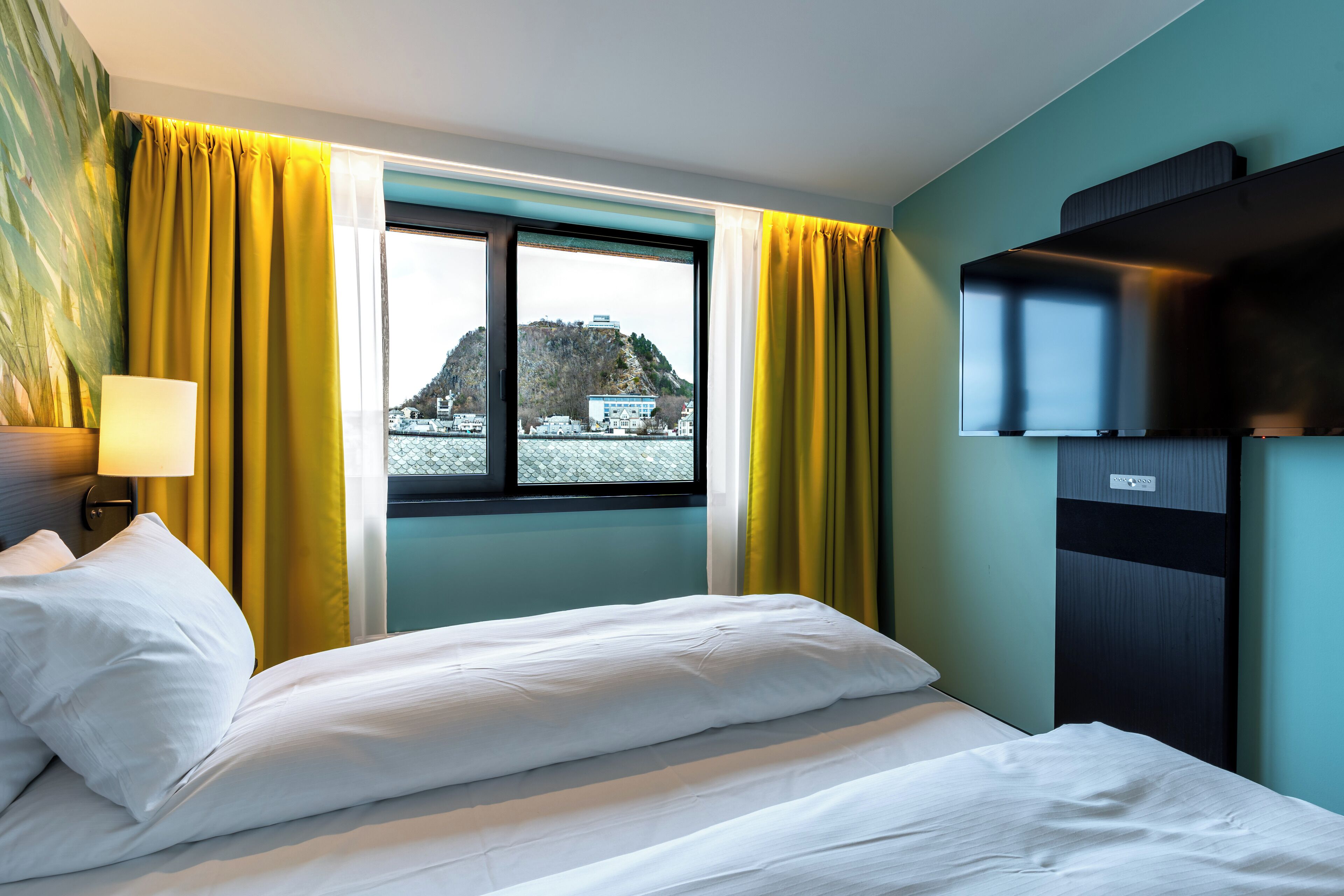 A hotel room with a bed, a window with curtains, a wall-mounted TV, and a view of a mountain.