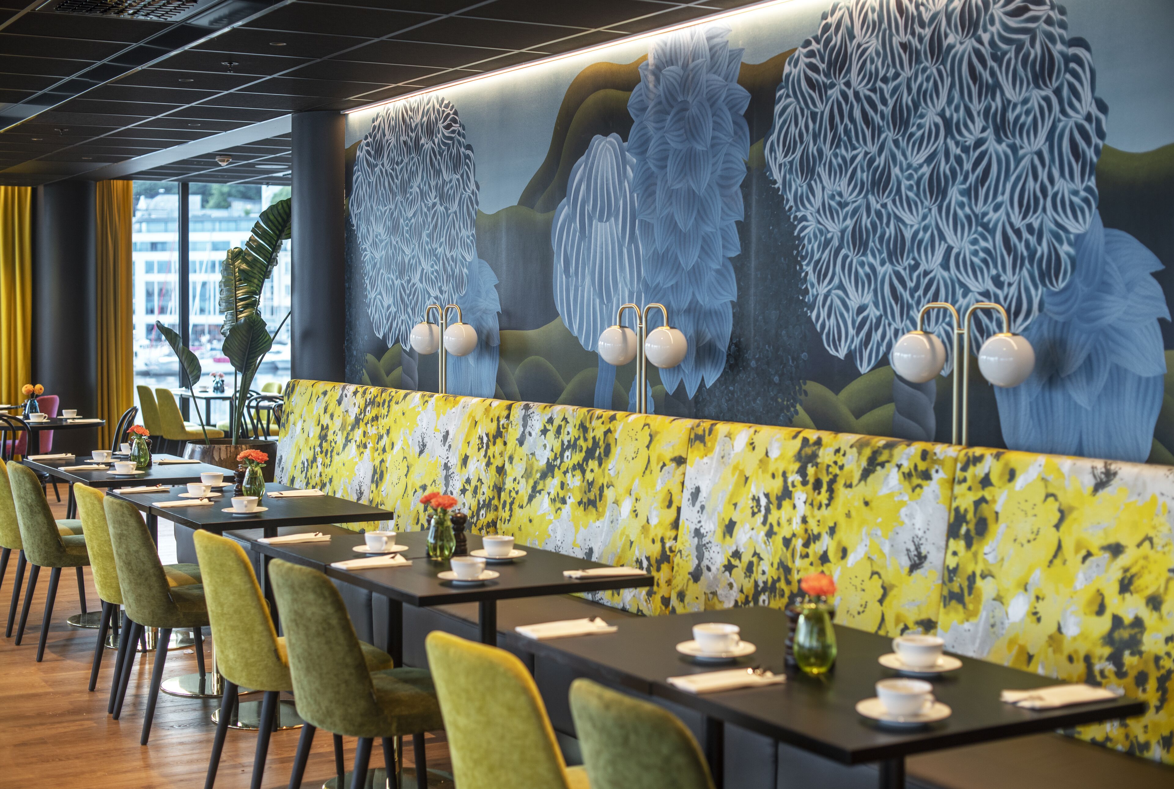 A restaurant with long tables, green chairs, and a yellow floral wall.