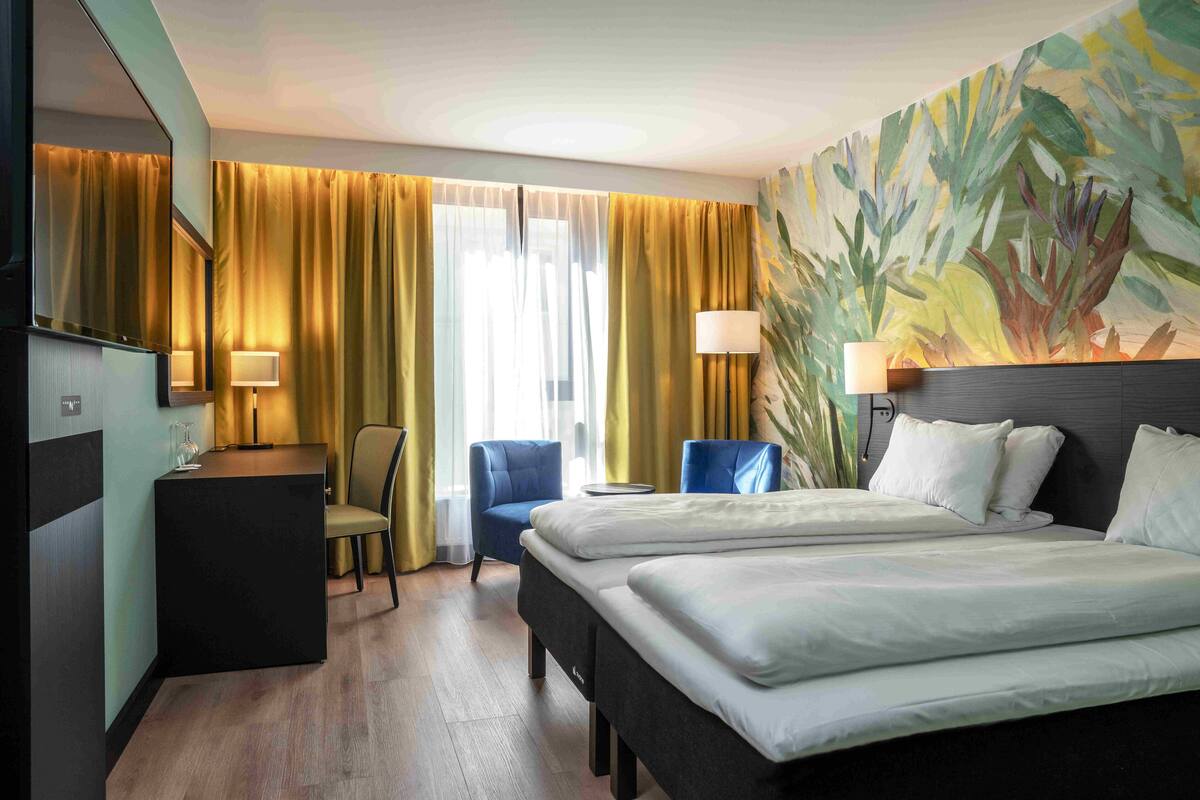 Hotellihuone, jossa on sänky, kaksi tuolia, työpöytä ja suuri ikkuna, jossa on verhot.