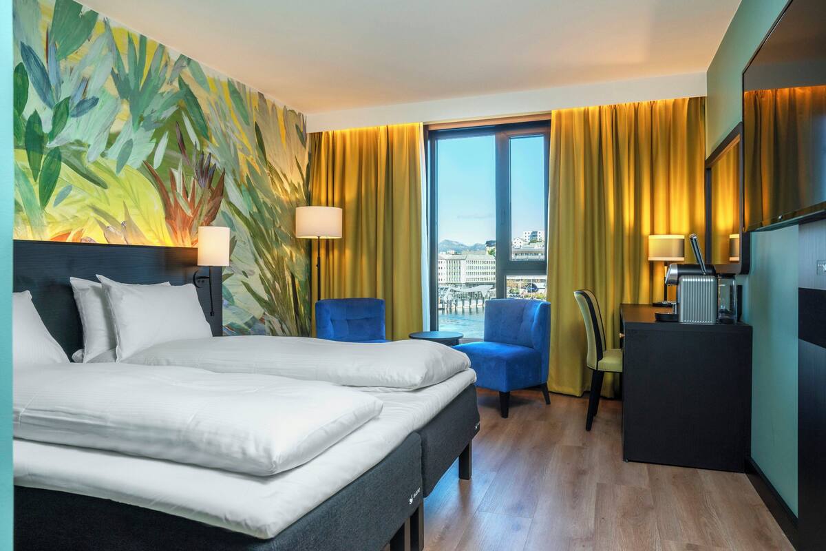 Hotellihuone, jossa on sänky, kaksi tuolia, työpöytä ja suuri ikkuna, jossa on verhot.