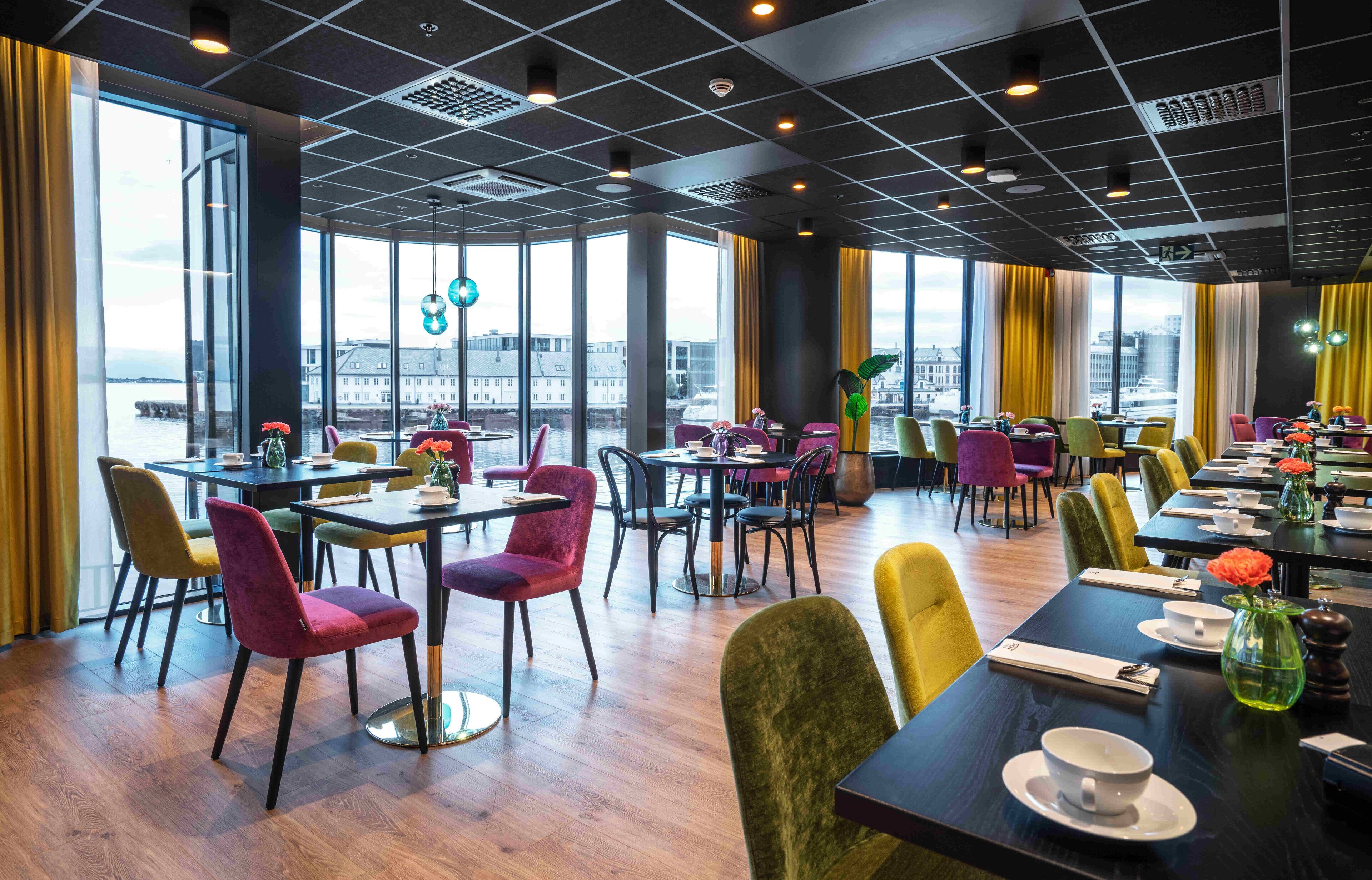 A modern dining area with multiple tables and chairs, large windows with a view of the city, and a high ceiling with grid lighting.