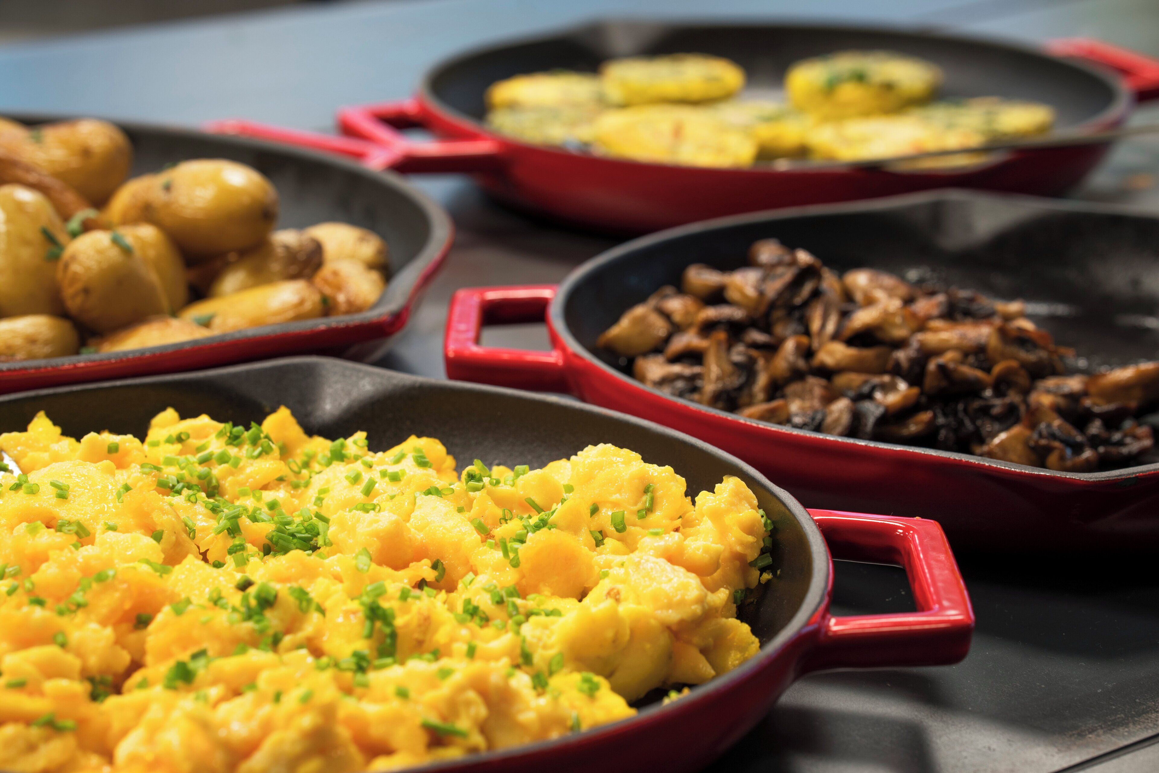 Trois poêles rouges contenant différents plats : des œufs brouillés, des pommes de terre rôties et des champignons sautés.