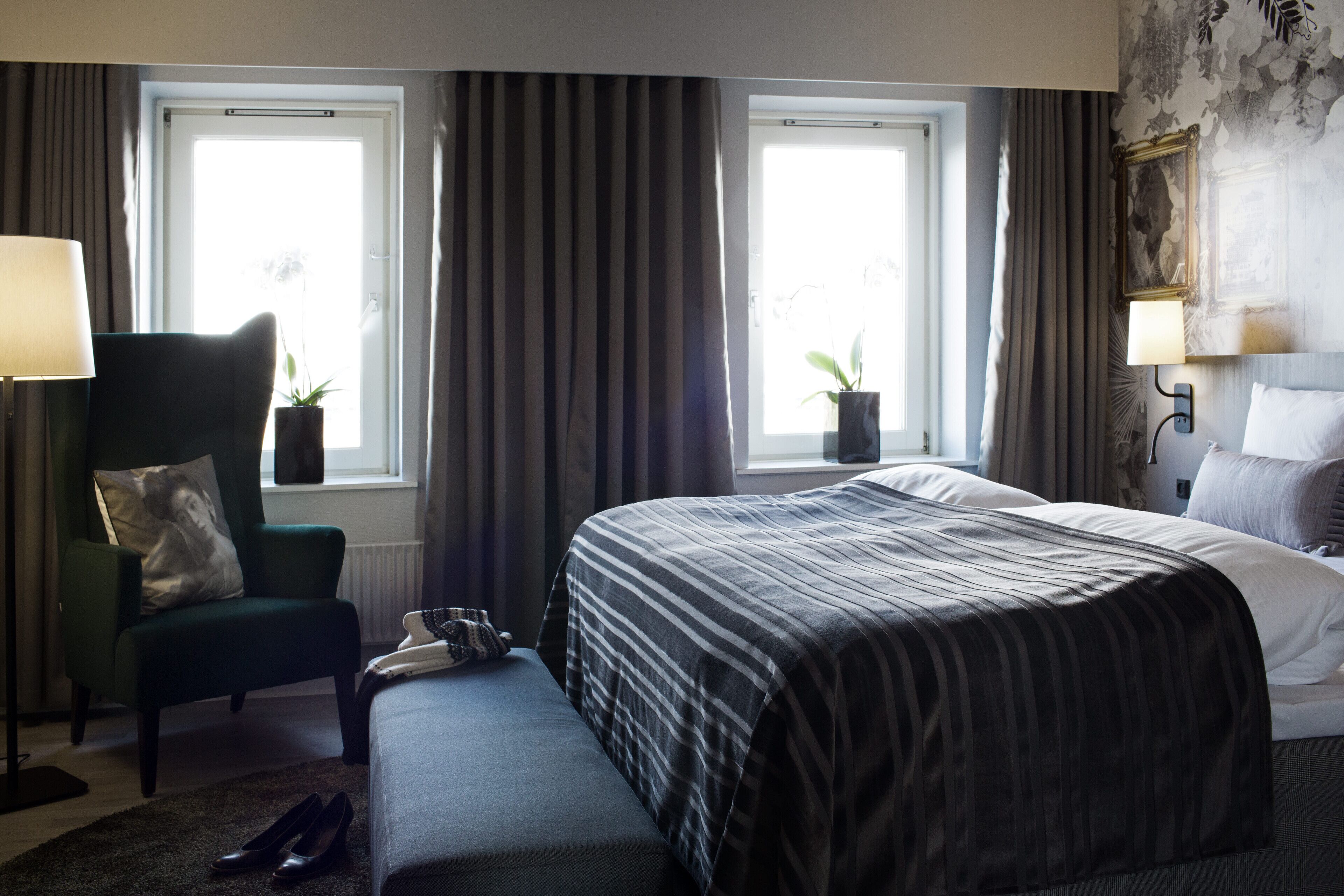 Une chambre d’hôtel avec un grand lit, une chaise, un banc, deux fenêtres avec des rideaux et une lampe fixée au mur.