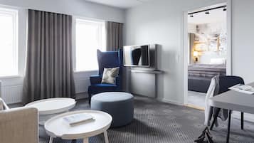Una habitación de hotel moderna con alfombra gris, un sillón azul, una mesa redonda blanca, un reposapiés gris, un televisor de pantalla plana y una cama con cabecera gris.