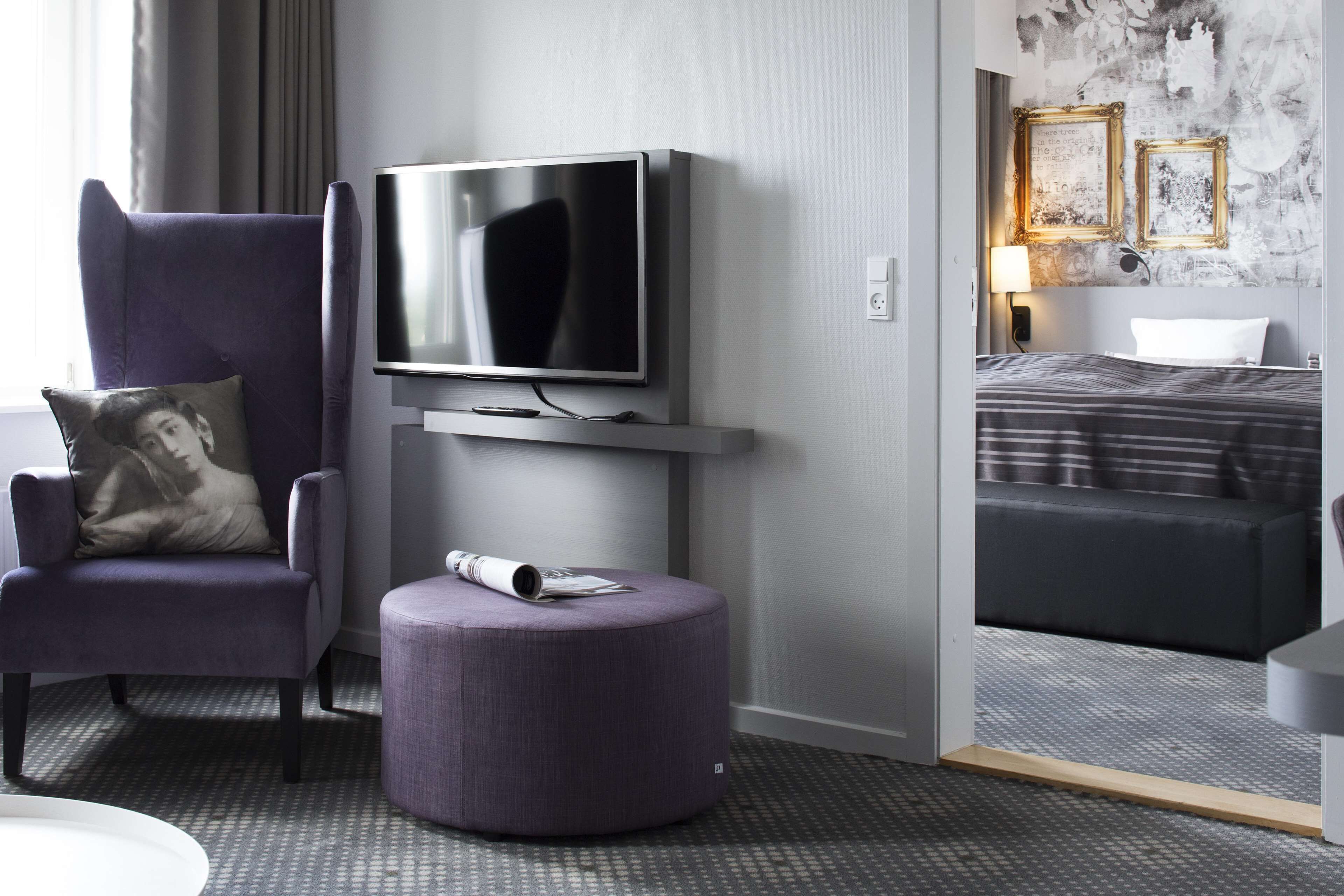 A modern hotel room with a flat-screen TV mounted on a wall, a comfortable armchair with a cushion, a round ottoman with a magazine, and a neatly made bed with a striped blanket.