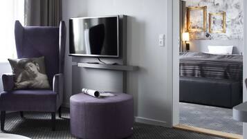 Una habitación de hotel moderna con un televisor de pantalla plana montado en la pared, un sillón cómodo con cojín, un otoman redondo con una revista, y una cama bien hecha con un edredón a rayas.