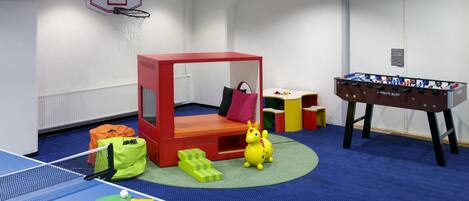 Una habitación con mesa de ping pong, mesa de futbolín, una cama pequeña y aro de baloncesto.