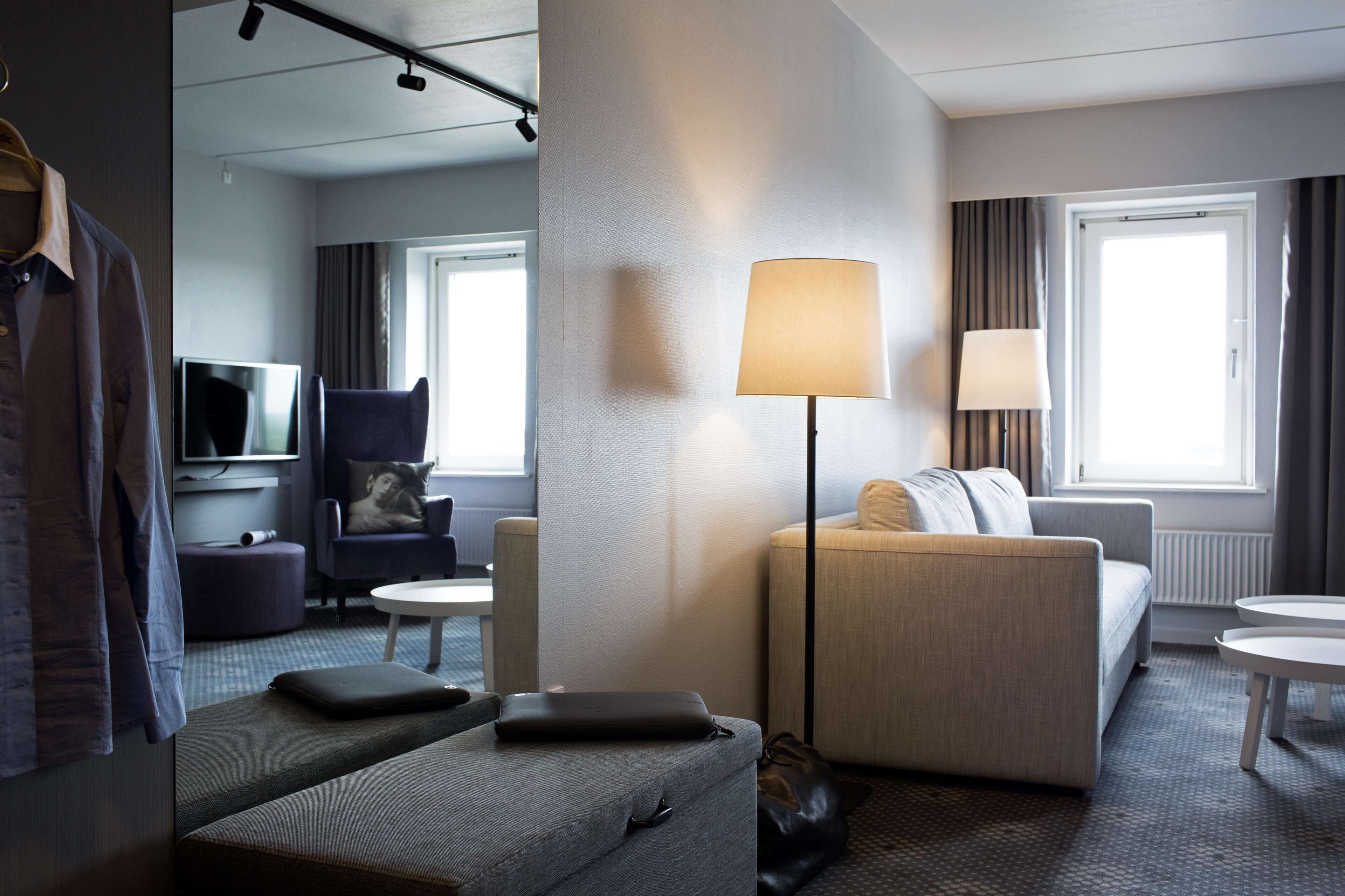 Ein modernes Hotelzimmer mit grauem Sofa, einem kleinen runden Tisch, einer Lampe, einem Fernseher und einem Kleiderhaken.