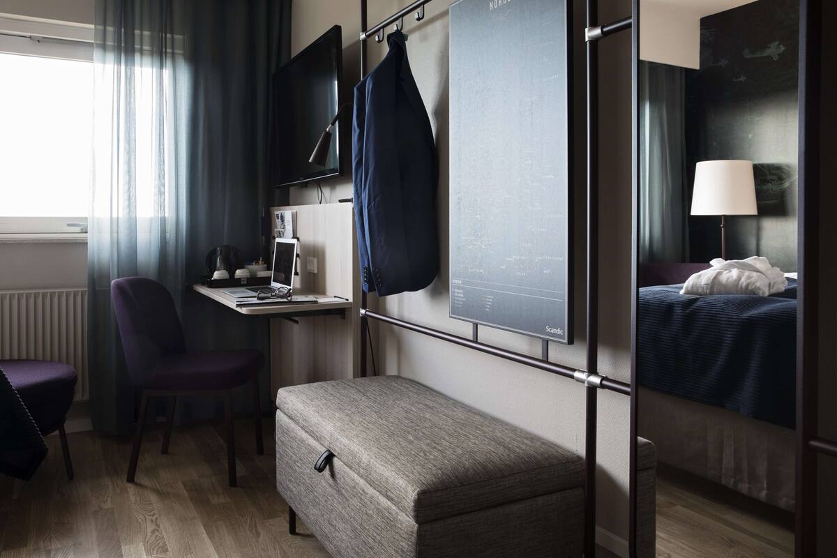 Hotellihuone, jossa on sänky, työpöytä tietokoneella, tuoli, penkki, televisio ja ikkuna, jossa on verhot.
