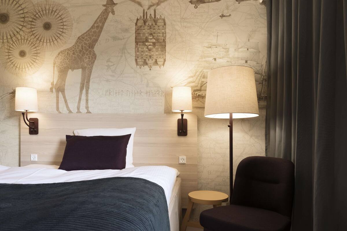 Hotellihuone, jossa on sänky, kaksi seinälle asennettua lamppua, karttateemalla koristeltu seinä ja tuoli.