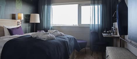 A hotel room with a large bed, a desk, a chair, a window with curtains, and a wall-mounted TV.
