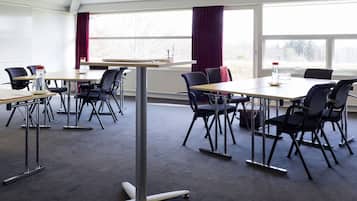 Un salón de conferencias con una gran mesa y varias sillas, ventanas amplias con cortinas y un radiador debajo de la mesa.
